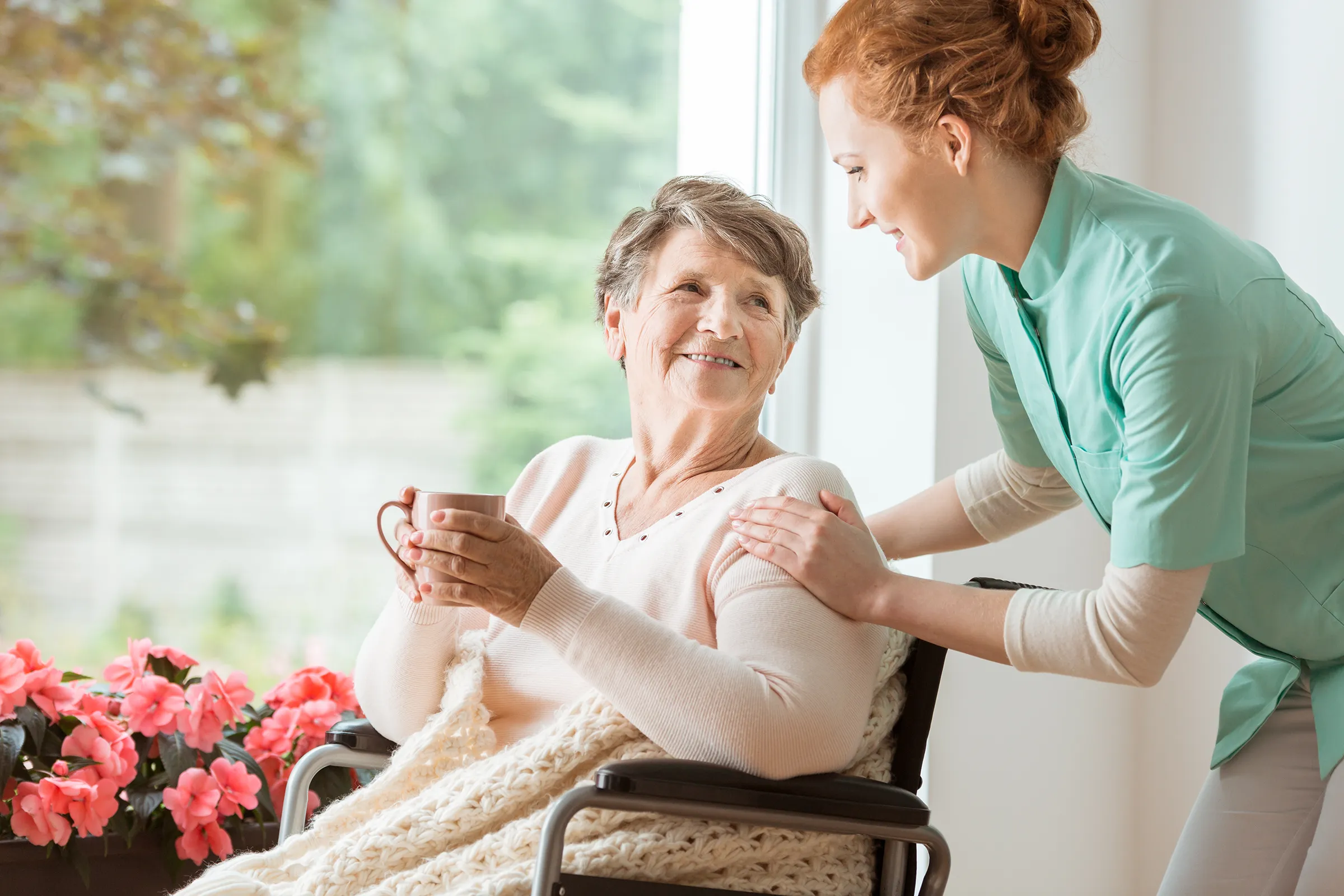 Caregiver assisting elderly woman with tea by window